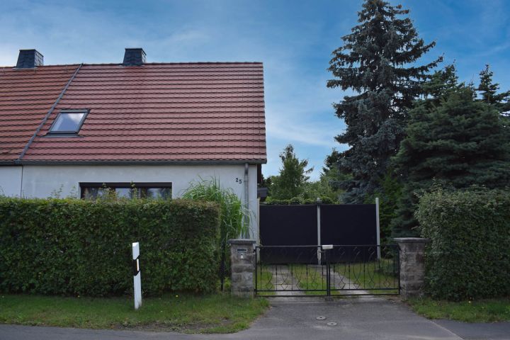Halbes Haus mit rotem Dach, Hecke, Tor und Bäumen im Hintergrund.