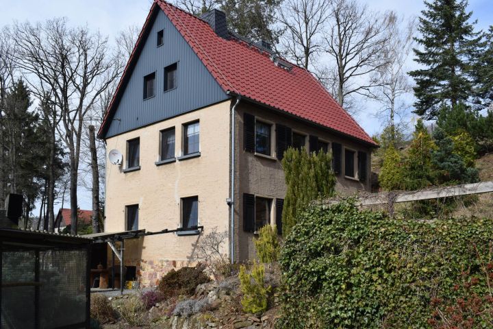 Ein zweistöckiges Haus mit rotem Dach, umgeben von Bäumen und Sträuchern im Garten.
