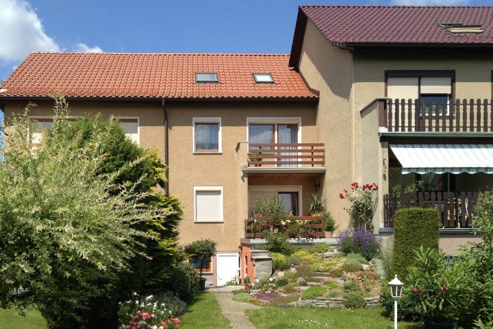 Zweistöckiges Haus mit Garten, roten Dachziegeln und Balkon, umgeben von Pflanzen.